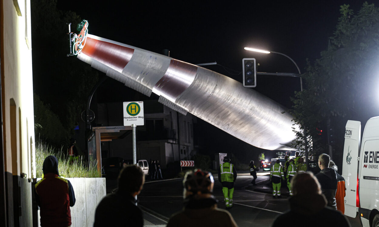Bei Nacht wird ein großes Rotorblatt einer Windkraftanlage mit einem Kran über eine Straße transportiert, während mehrere Menschen und Einsatzkräfte den Transport beobachten.