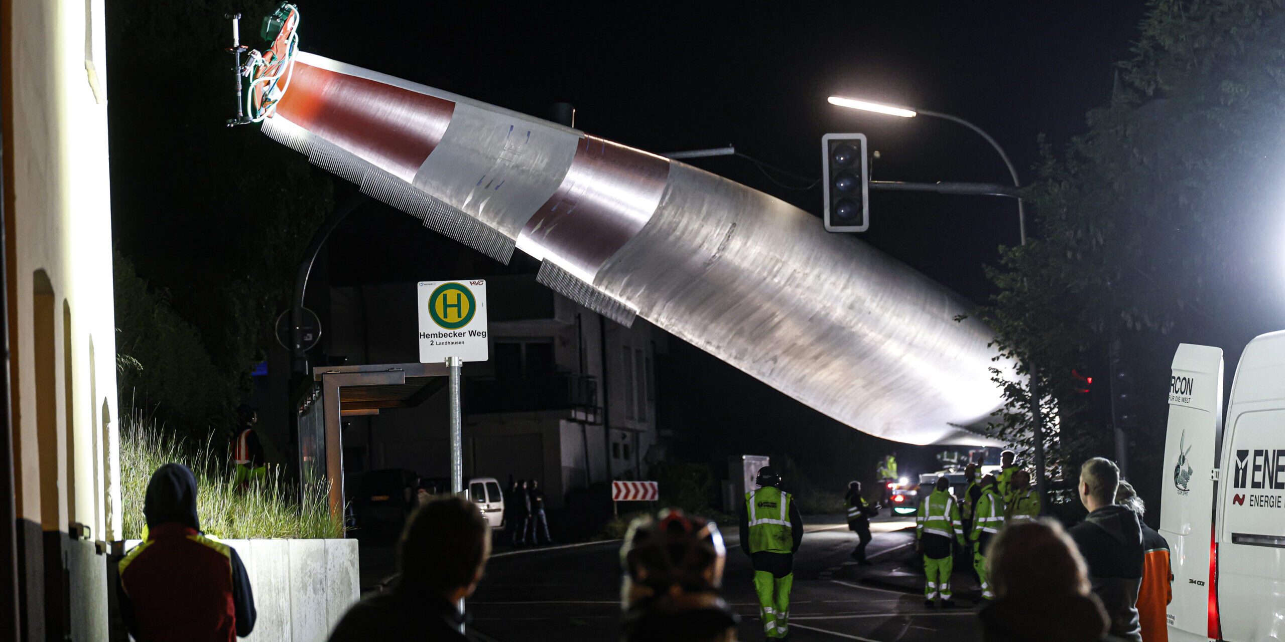 Bei Nacht wird ein großes Rotorblatt einer Windkraftanlage mit einem Kran über eine Straße transportiert, während mehrere Menschen und Einsatzkräfte den Transport beobachten.