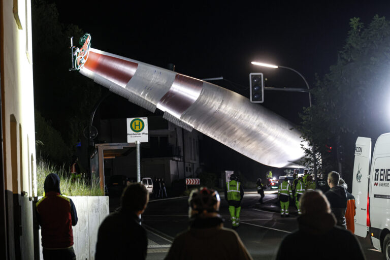 Bei Nacht wird ein großes Rotorblatt einer Windkraftanlage mit einem Kran über eine Straße transportiert, während mehrere Menschen und Einsatzkräfte den Transport beobachten.
