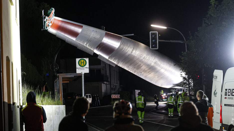 Bei Nacht wird ein großes Rotorblatt einer Windkraftanlage mit einem Kran über eine Straße transportiert, während mehrere Menschen und Einsatzkräfte den Transport beobachten.