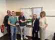 Fünf Personen stehen neben einem neu installierten Defibrillator und einem Erste-Hilfe-Kasten an einer Wand mit Flucht- und Rettungsplan.