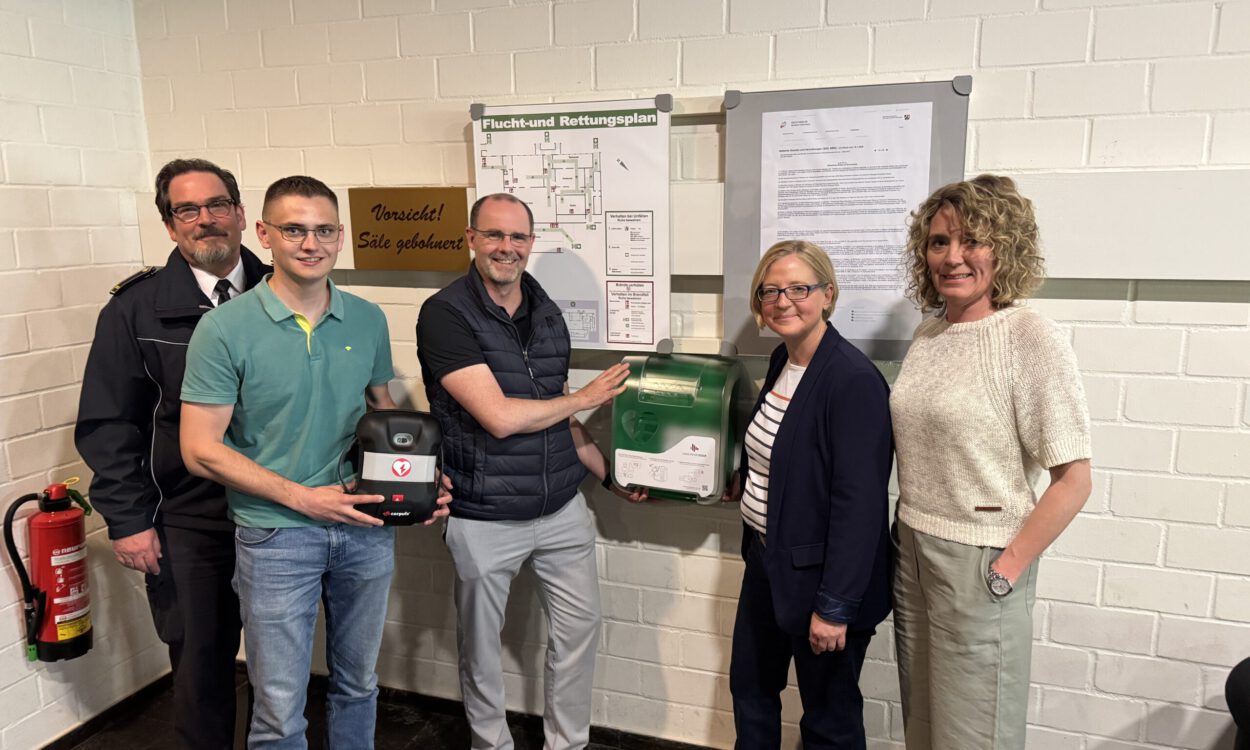 Fünf Personen stehen neben einem neu installierten Defibrillator und einem Erste-Hilfe-Kasten an einer Wand mit Flucht- und Rettungsplan.