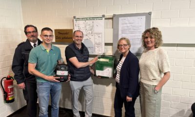 Fünf Personen stehen neben einem neu installierten Defibrillator und einem Erste-Hilfe-Kasten an einer Wand mit Flucht- und Rettungsplan.
