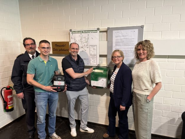 Foto: Stadt Menden Fünf Personen stehen neben einem neu installierten Defibrillator und einem Erste-Hilfe-Kasten an einer Wand mit Flucht- und Rettungsplan.