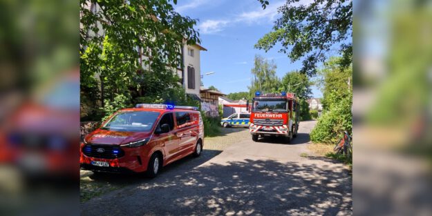 Mehrere Einsatzfahrzeuge von Feuerwehr und Polizei stehen an einer schmalen Straße zwischen Bäumen und Wohnhäusern.