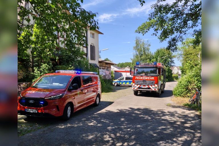 Mehrere Einsatzfahrzeuge von Feuerwehr und Polizei stehen an einer schmalen Straße zwischen Bäumen und Wohnhäusern.