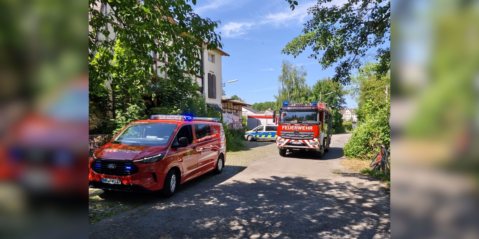 Mehrere Einsatzfahrzeuge von Feuerwehr und Polizei stehen an einer schmalen Straße zwischen Bäumen und Wohnhäusern.