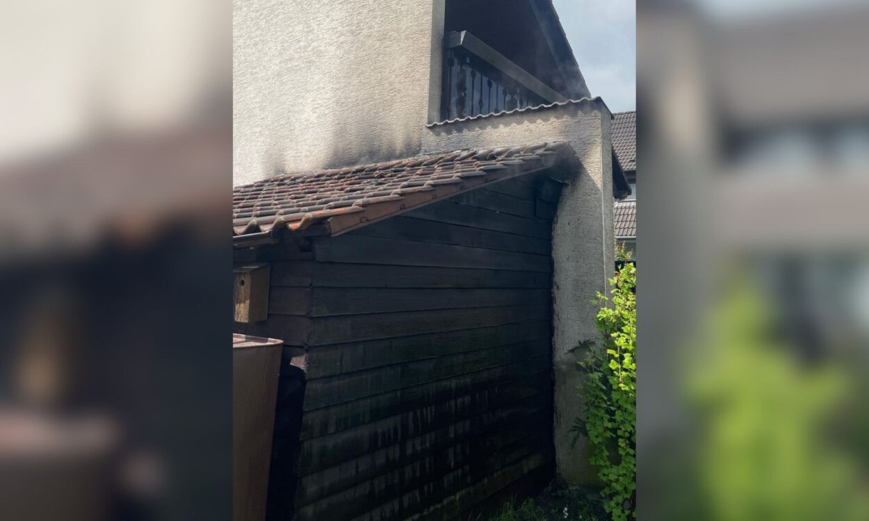 An einem Wohnhaus sind deutliche Rußspuren an einer angebauten Holzhütte und am Dachbereich nach einem Brand zu sehen.