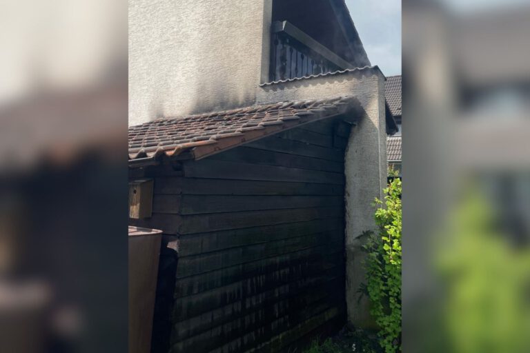 An einem Wohnhaus sind deutliche Rußspuren an einer angebauten Holzhütte und am Dachbereich nach einem Brand zu sehen.