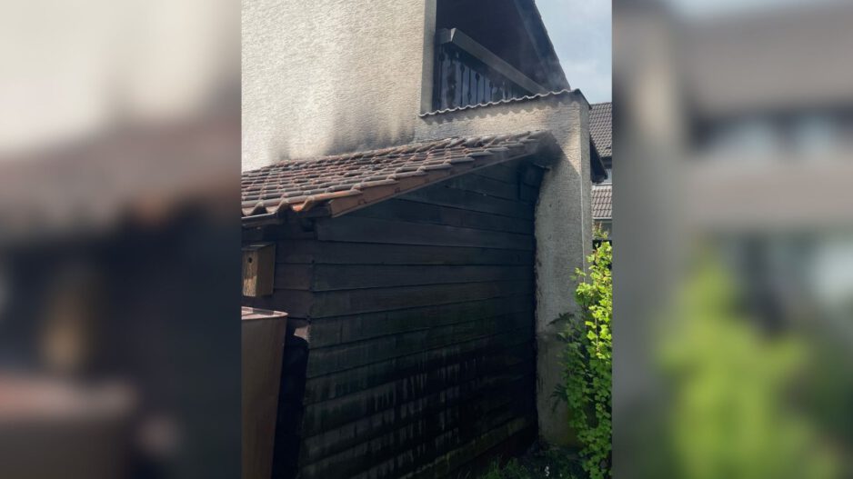 An einem Wohnhaus sind deutliche Rußspuren an einer angebauten Holzhütte und am Dachbereich nach einem Brand zu sehen.