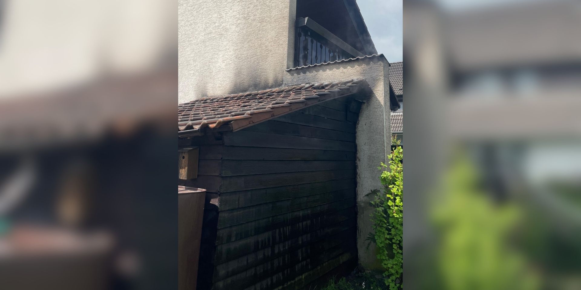 An einem Wohnhaus sind deutliche Rußspuren an einer angebauten Holzhütte und am Dachbereich nach einem Brand zu sehen.