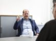 Ein Mann im Sakko sitzt in einem Büro an einem Tisch und gestikuliert während eines Gesprächs mit einer gegenüber sitzenden Person, vor ihm steht eine Kaffeetasse.