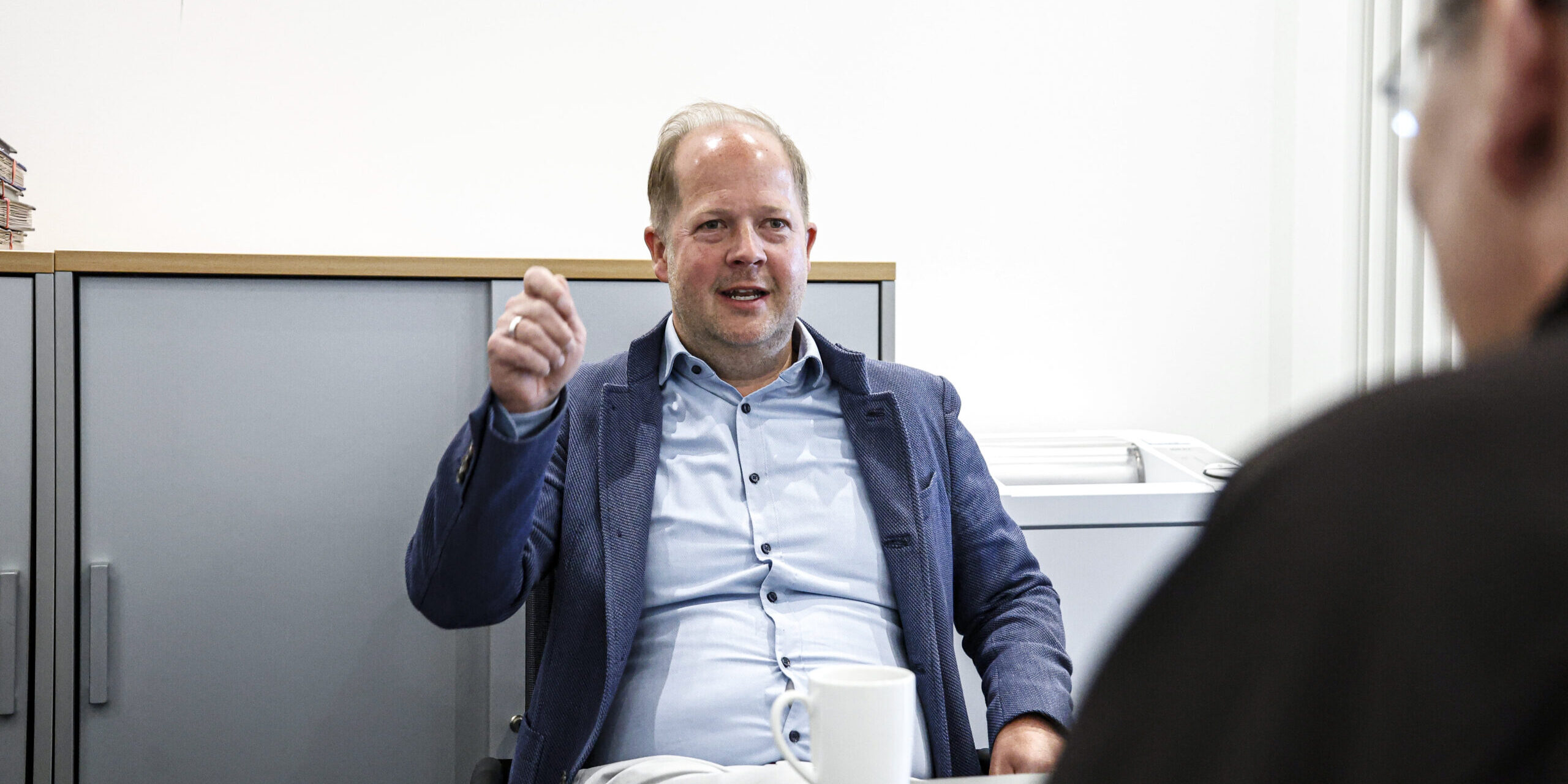 Ein Mann im Sakko sitzt in einem Büro an einem Tisch und gestikuliert während eines Gesprächs mit einer gegenüber sitzenden Person, vor ihm steht eine Kaffeetasse.