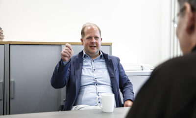 Ein Mann im Sakko sitzt in einem Büro an einem Tisch und gestikuliert während eines Gesprächs mit einer gegenüber sitzenden Person, vor ihm steht eine Kaffeetasse.