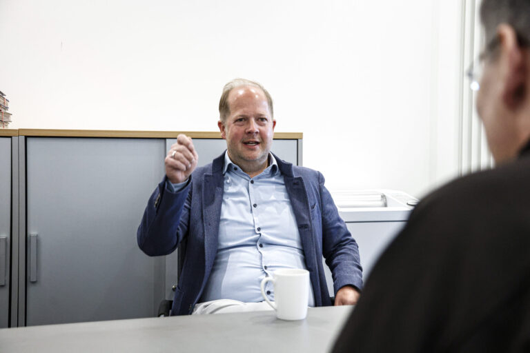 Ein Mann im Sakko sitzt in einem Büro an einem Tisch und gestikuliert während eines Gesprächs mit einer gegenüber sitzenden Person, vor ihm steht eine Kaffeetasse.