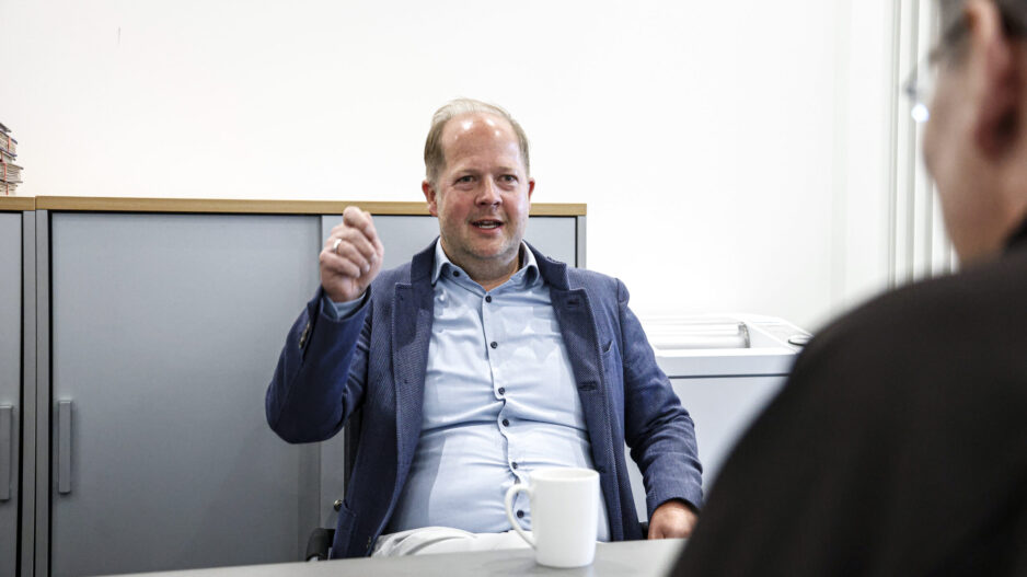 Ein Mann im Sakko sitzt in einem Büro an einem Tisch und gestikuliert während eines Gesprächs mit einer gegenüber sitzenden Person, vor ihm steht eine Kaffeetasse.