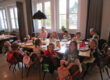 Mehrere Kinder sitzen gemeinsam mit Betreuerinnen an einem gedeckten Tisch in einem hellen Gruppenraum einer Kita.
