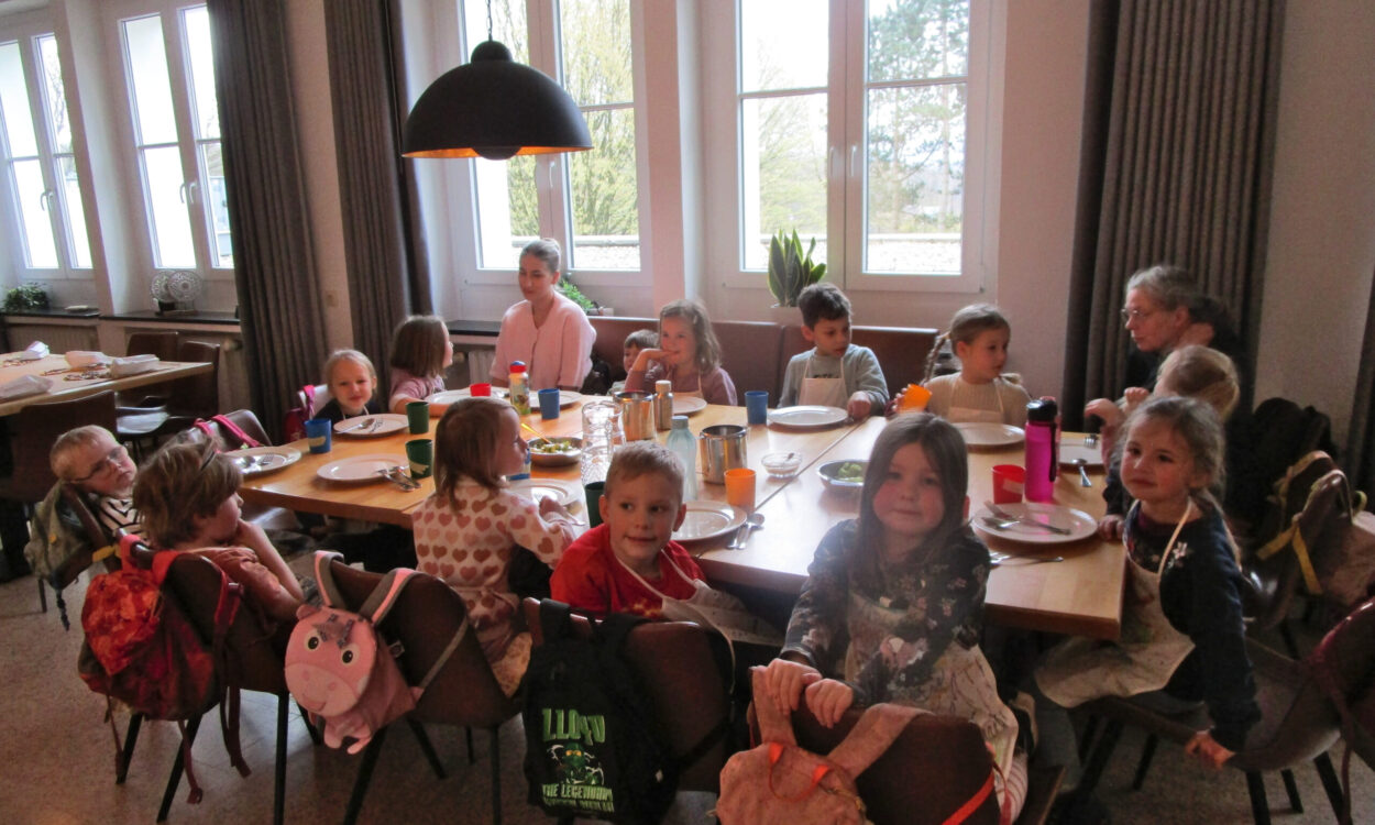 Mehrere Kinder sitzen gemeinsam mit Betreuerinnen an einem gedeckten Tisch in einem hellen Gruppenraum einer Kita.