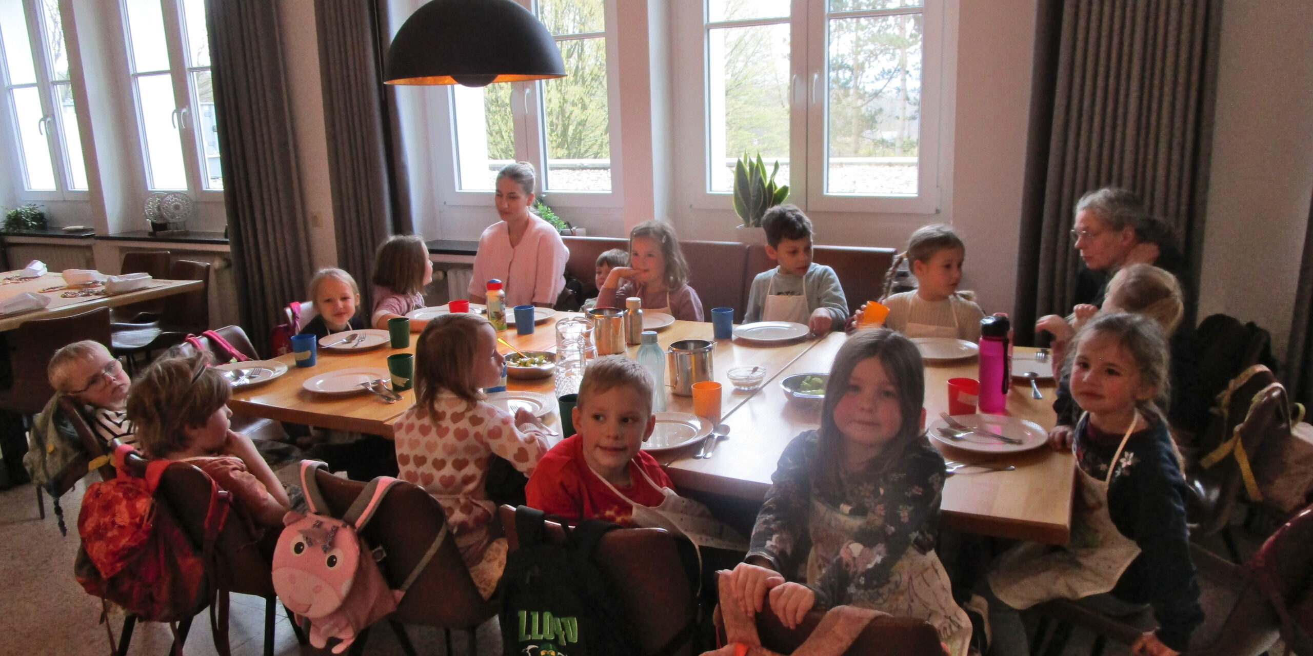Mehrere Kinder sitzen gemeinsam mit Betreuerinnen an einem gedeckten Tisch in einem hellen Gruppenraum einer Kita.