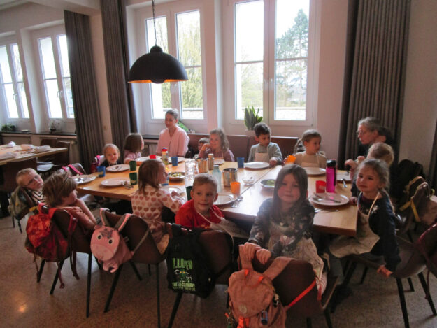 Foto: Stadt Menden Mehrere Kinder sitzen gemeinsam mit Betreuerinnen an einem gedeckten Tisch in einem hellen Gruppenraum einer Kita.
