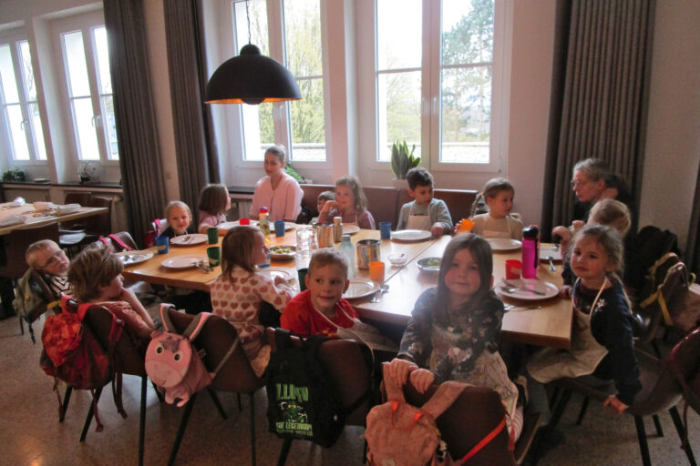 Mehrere Kinder sitzen gemeinsam mit Betreuerinnen an einem gedeckten Tisch in einem hellen Gruppenraum einer Kita.