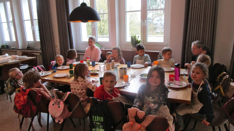 Mehrere Kinder sitzen gemeinsam mit Betreuerinnen an einem gedeckten Tisch in einem hellen Gruppenraum einer Kita.
