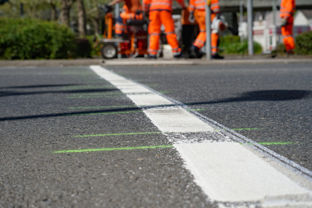 Frisch aufgebrachte weiße Straßenmarkierung auf Asphalt im Vordergrund, während im Hintergrund unscharf Straßenarbeiter in orangefarbener Schutzkleidung arbeiten.