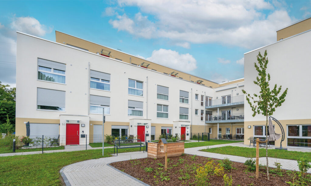 Ein modernes Wohngebäude mit heller Fassade und roten Eingangstüren um einen begrünten Innenhof mit Wegen, Beeten und Sitzmöglichkeiten.