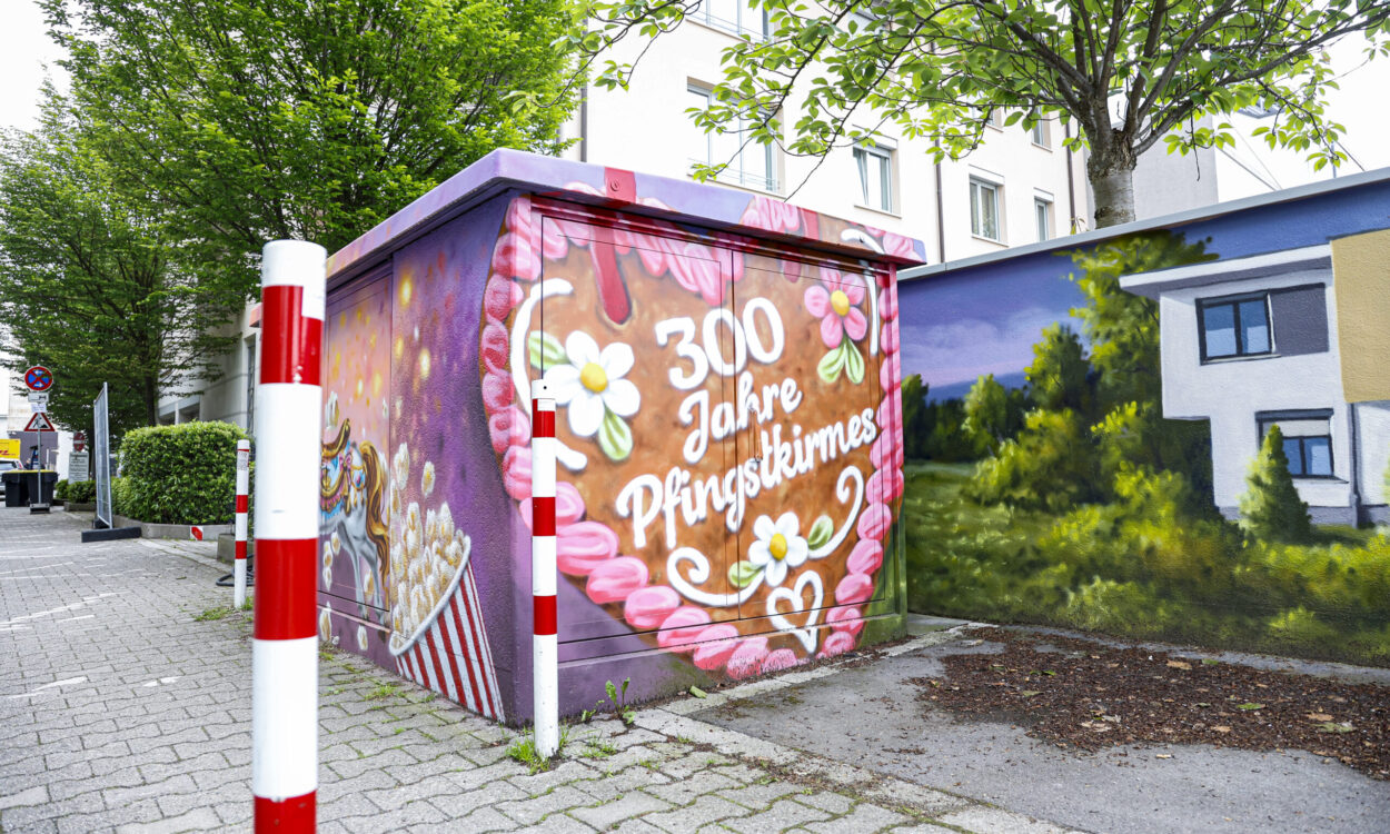 Ein bunt bemaltes Trafohäuschen mit der Aufschrift „300 Jahre Pfingstkirmes“ steht an einem Gehweg zwischen Bäumen und Wohnhäusern.
