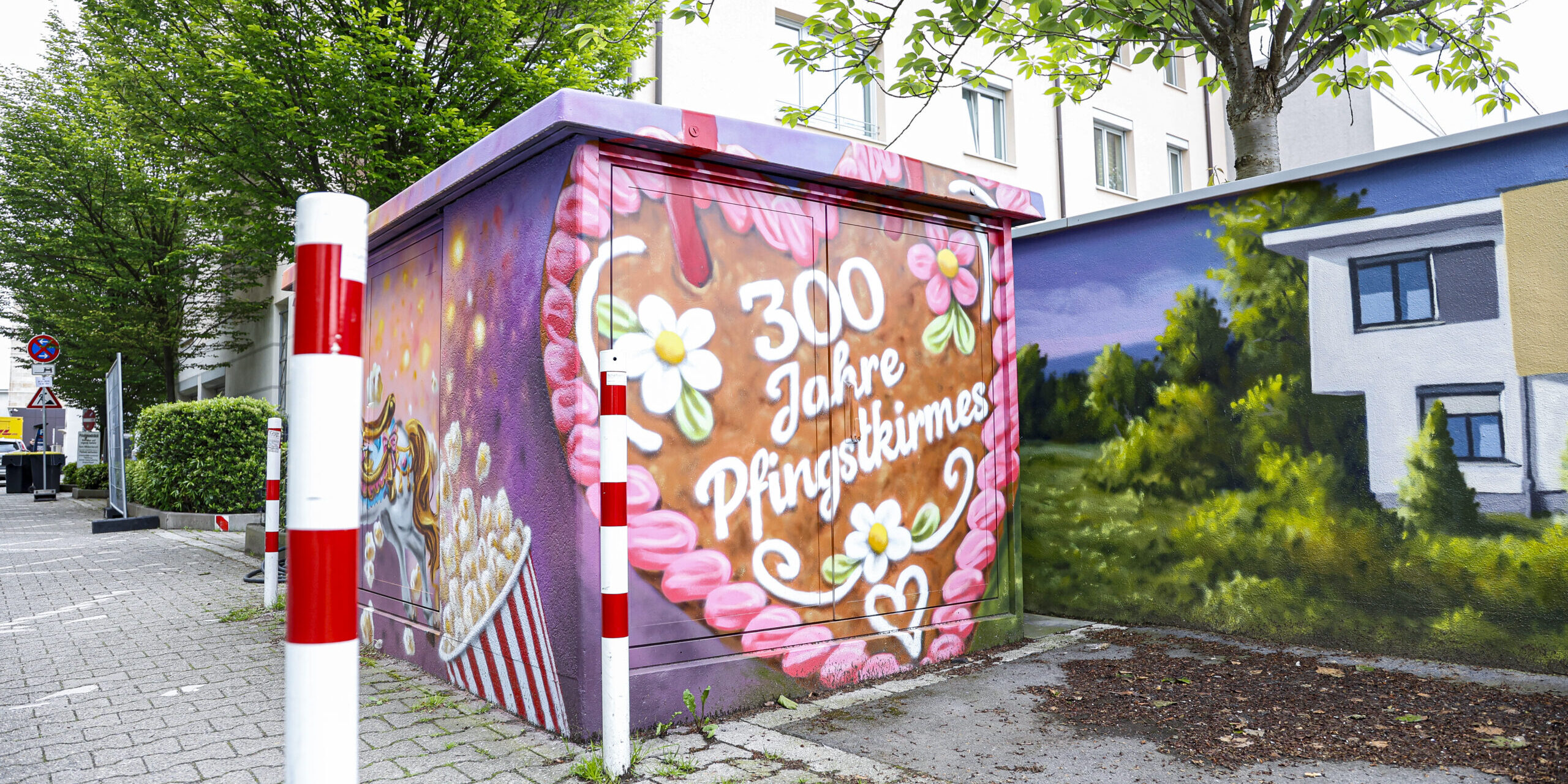 Ein bunt bemaltes Trafohäuschen mit der Aufschrift „300 Jahre Pfingstkirmes“ steht an einem Gehweg zwischen Bäumen und Wohnhäusern.