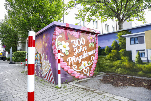 Foto: Lucas Sellmann Ein bunt bemaltes Trafohäuschen mit der Aufschrift „300 Jahre Pfingstkirmes“ steht an einem Gehweg zwischen Bäumen und Wohnhäusern.