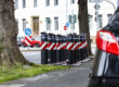 Mehrere schwarz-rote Absperrpoller stehen am Straßenrand zwischen Bäumen an einer Kreuzung in einem Wohngebiet.