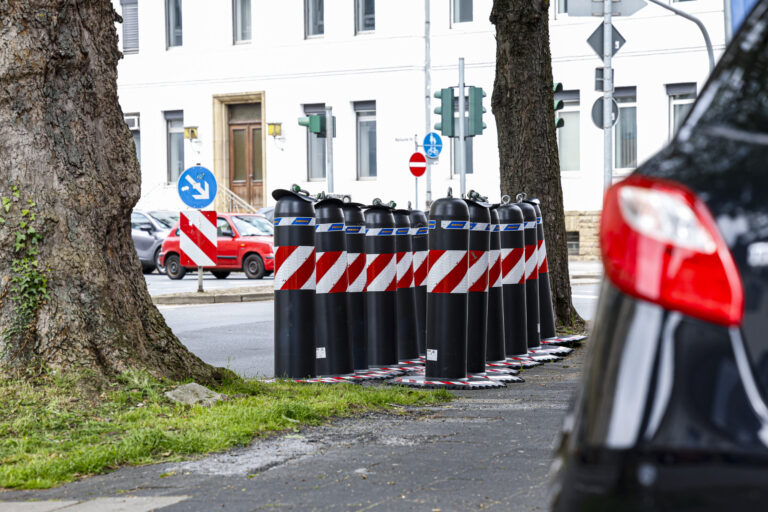 Mehrere schwarz-rote Absperrpoller stehen am Straßenrand zwischen Bäumen an einer Kreuzung in einem Wohngebiet.