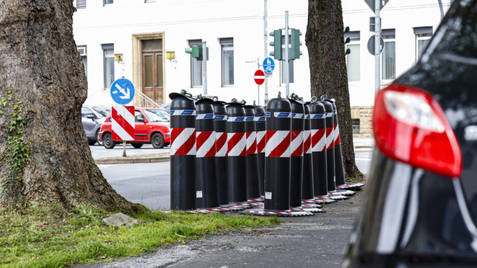 Mehrere schwarz-rote Absperrpoller stehen am Straßenrand zwischen Bäumen an einer Kreuzung in einem Wohngebiet.