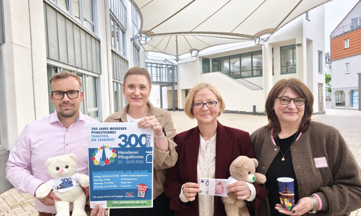 Vier Personen präsentieren ein Plakat und Werbeartikel zum 300-jährigen Jubiläum der Mendener Pfingstkirmes in einem überdachten Innenhof.