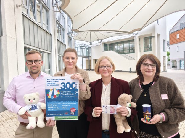 Foto: Stadt Menden/Denise Rüscher Vier Personen präsentieren ein Plakat und Werbeartikel zum 300-jährigen Jubiläum der Mendener Pfingstkirmes in einem überdachten Innenhof.