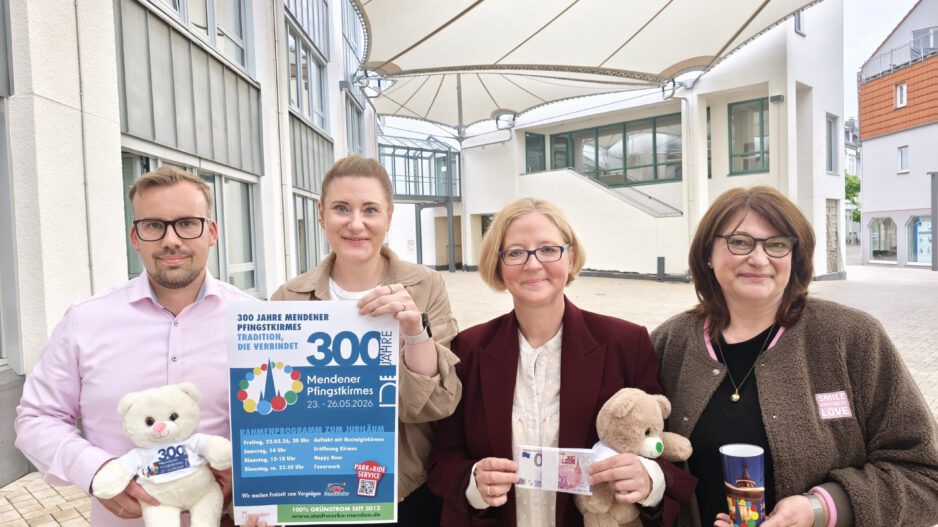 Vier Personen präsentieren ein Plakat und Werbeartikel zum 300-jährigen Jubiläum der Mendener Pfingstkirmes in einem überdachten Innenhof.