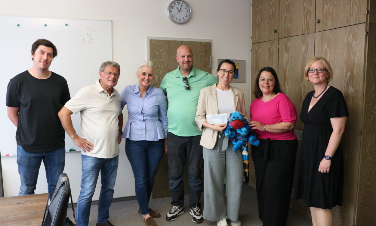 Sieben Personen stehen in einem Besprechungsraum für ein Gruppenfoto zusammen. Eine Frau hält einen symbolischen Spendenscheck, daneben ist ein blaues Kuscheltier zu sehen.