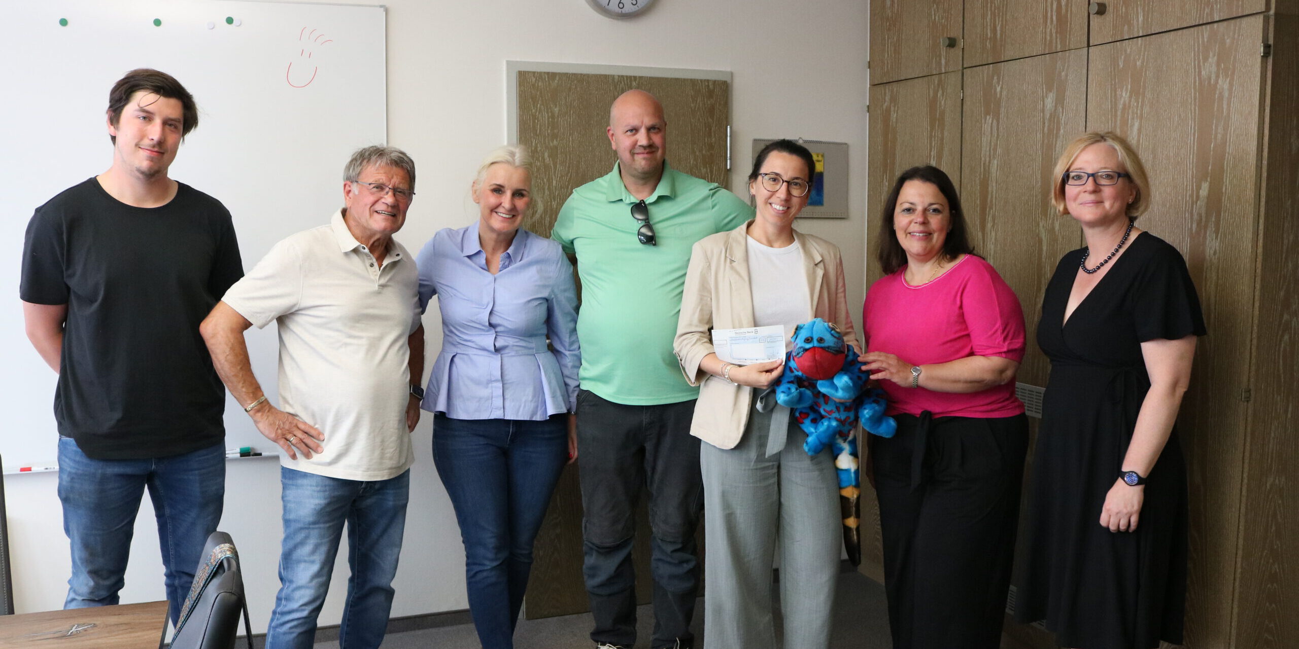 Sieben Personen stehen in einem Besprechungsraum für ein Gruppenfoto zusammen. Eine Frau hält einen symbolischen Spendenscheck, daneben ist ein blaues Kuscheltier zu sehen.