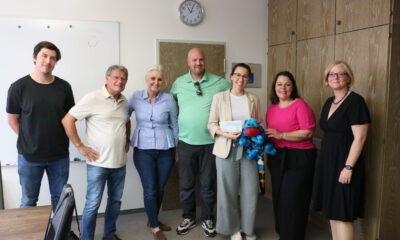 Sieben Personen stehen in einem Besprechungsraum für ein Gruppenfoto zusammen. Eine Frau hält einen symbolischen Spendenscheck, daneben ist ein blaues Kuscheltier zu sehen.