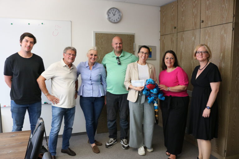 Sieben Personen stehen in einem Besprechungsraum für ein Gruppenfoto zusammen. Eine Frau hält einen symbolischen Spendenscheck, daneben ist ein blaues Kuscheltier zu sehen.