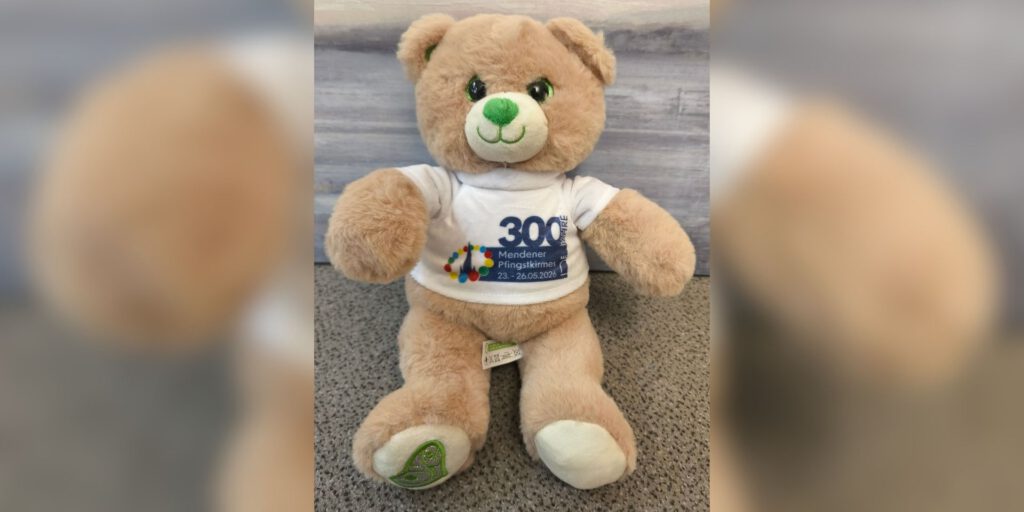 Ein heller Plüschteddy mit grünem Herz auf der Pfote trägt ein T-Shirt zum 300-jährigen Jubiläum der Mendener Pfingstkirmes 2026.