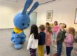 Mehrere Kinder stehen in einem Raum und begegnen einer großen blauen Hasenfigur, die sich zu ihnen hinunterbeugt und mit ihnen interagiert.