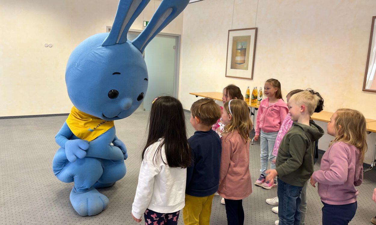 Mehrere Kinder stehen in einem Raum und begegnen einer großen blauen Hasenfigur, die sich zu ihnen hinunterbeugt und mit ihnen interagiert.