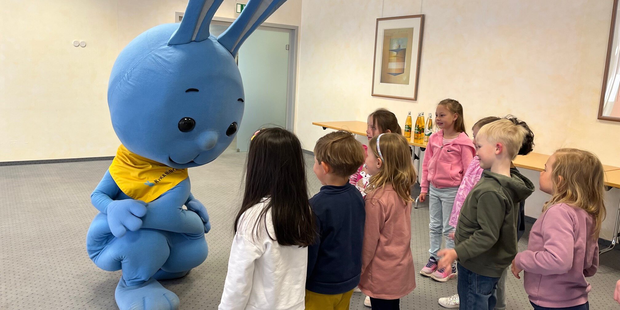 Mehrere Kinder stehen in einem Raum und begegnen einer großen blauen Hasenfigur, die sich zu ihnen hinunterbeugt und mit ihnen interagiert.