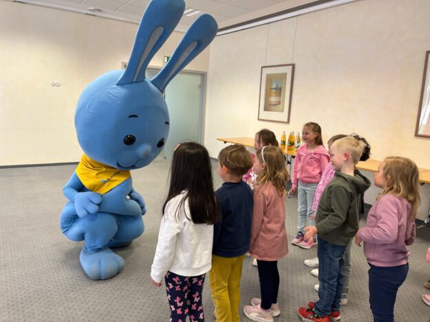 Mehrere Kinder stehen in einem Raum und begegnen einer großen blauen Hasenfigur, die sich zu ihnen hinunterbeugt und mit ihnen interagiert.