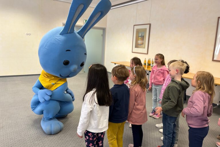 Mehrere Kinder stehen in einem Raum und begegnen einer großen blauen Hasenfigur, die sich zu ihnen hinunterbeugt und mit ihnen interagiert.