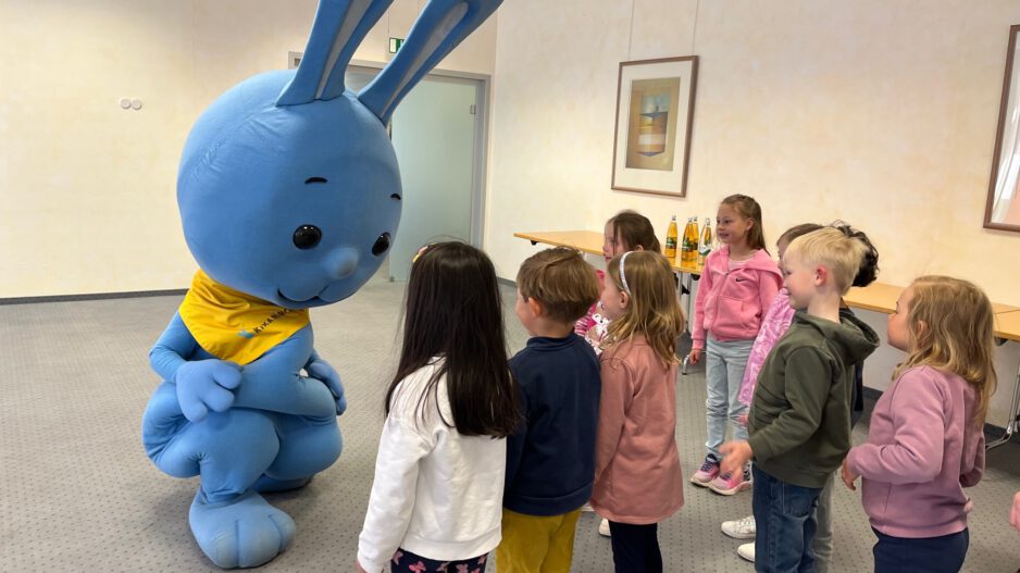 Mehrere Kinder stehen in einem Raum und begegnen einer großen blauen Hasenfigur, die sich zu ihnen hinunterbeugt und mit ihnen interagiert.