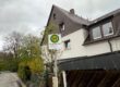 Ein Haltestellenschild mit der Aufschrift „Waldecker Hof“ steht vor einem Wohnhaus an einer ruhigen Straße, umgeben von Bäumen und Hecken unter grauem Himmel.