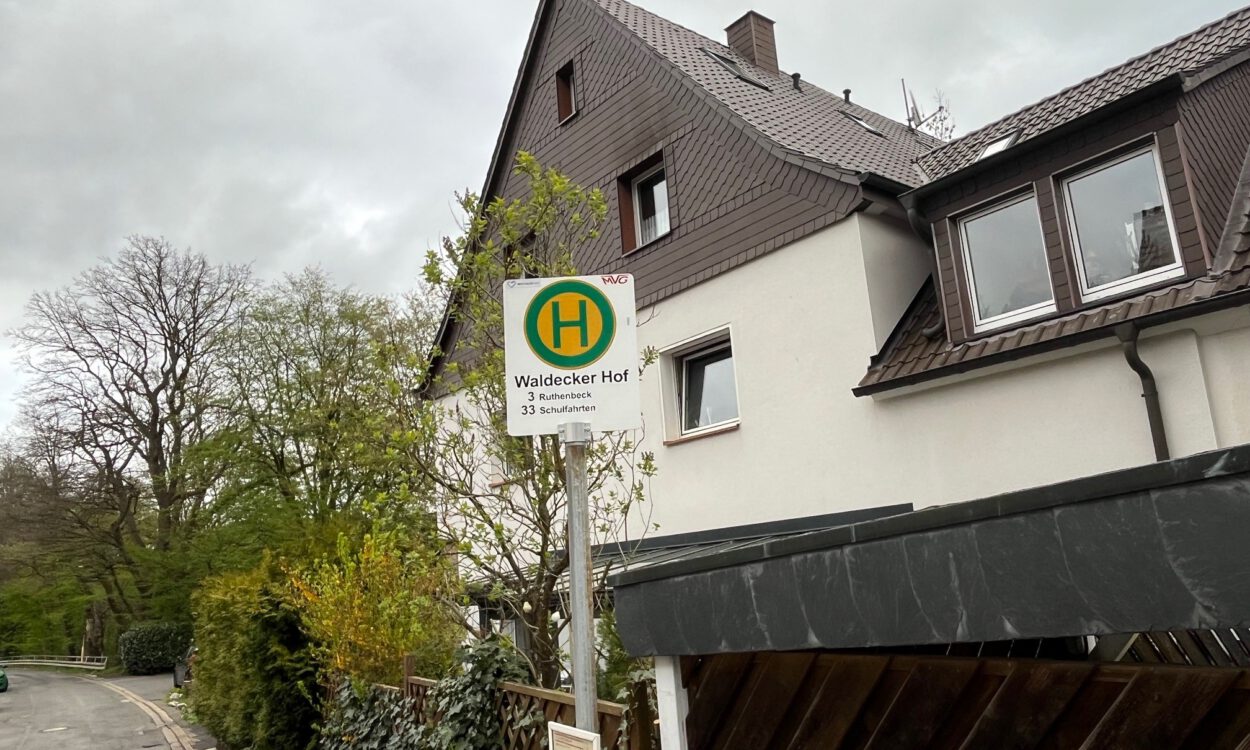 Ein Haltestellenschild mit der Aufschrift „Waldecker Hof“ steht vor einem Wohnhaus an einer ruhigen Straße, umgeben von Bäumen und Hecken unter grauem Himmel.
